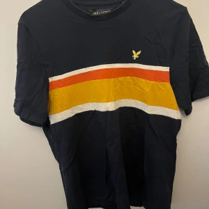 Randig t-shirt från Lyle & Scott - Snygg t-shirt från Lyle & Scott med marinblå bas och breda ränder i vitt, gult och rött över bröstet. Klassisk rund hals och korta ärmar. Liten gul logga broderad på vänster sida. Mjuk bomullskvalitet som är skön mot huden.