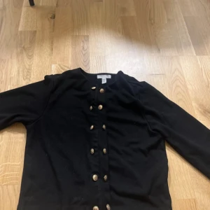 Svart kofta med guldknappar - Svart kofta från H&M med snygga guldknappar framtill. Klassisk rund halsringning och långärmad modell. Perfekt att slänga på sig när det är lite kyligt. Materialet känns mjukt och skönt mot huden, troligtvis bomull eller en bomullsblandning. #hm