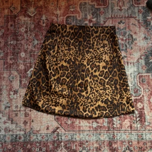 Leopardmönstrad kortkjol i satin - Snygg kortkjol med leopardmönster i bruna och svarta toner. Kjolen har en lätt A-linjeform och är gjord i ett glansigt satinmaterial som ger en cool och trendig vibe. Perfekt för dig som vill sticka ut med din outfit.