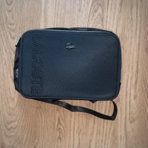 Svart axelväska från Lacoste - Snygg svart axelväska från Lacoste med tydlig logga och krokodil-emblem på framsidan. Väskan har en justerbar rem och är tillverkad i slitstark polyester. Perfekt storlek för det viktigaste och stilren design som passar till många outfits.