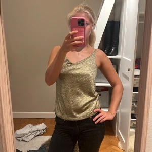 Glittrig guld linnetopp med omlott - Säljer en glittrig guld linnetopp med omlott-detalj framtill. Toppen har smala axelband och ärmlös design som ger en snygg och festlig look. Materialet är mjukt och skimrande, perfekt för att sticka ut. Passformen är normal och toppen är lätt att matcha med jeans eller kjol.