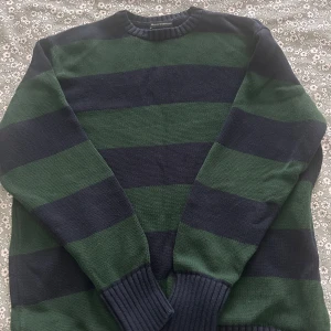 Randig stickad tröja Brandy Melville - Säljer en stickad tröja från Brandy Melville med breda marinblå och gröna ränder. Tröjan har rund hals, ribbade muddar och långa ärmar. Perfekt för dig som gillar chill och enkel stil med lite preppy känsla.