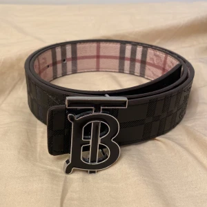 Burberry svart och rutig skärp - Snyggt Burberry-bälte i svart skinn med klassiskt rutmönster och präglad ryttare. Insidan har den ikoniska Burberry-rutan i beige, rosa och grått. Spännet är i silverfärgad metall med en stor B-logga. Perfekt accessoar för att lyfta din outfit.