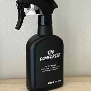 Lush The Comforter Body Spray 200ml - En svart sprayflaska med The Comforter Body Spray från Lush. Doften har inslag av svartvinbär och bergamott. Flaskan rymmer 200 ml och har en modern, stilren design med vit text på svart bakgrund. Köpt en av tjej här på plick men doften var tyvärr inte för mig