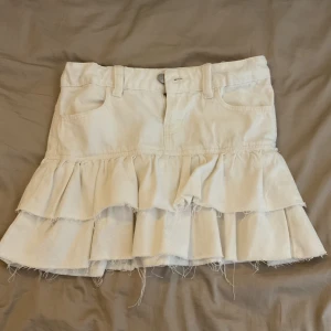 Vit volangkjol i denim från Gina Tricot - Supersöt vit jeanskjol med låg midja och dubbla volanger. Kjolen har klassiska fickor fram och bälteshällor, samt råa kanter för en trendig look. Perfekt för dig som gillar en lekfull och cool stil.