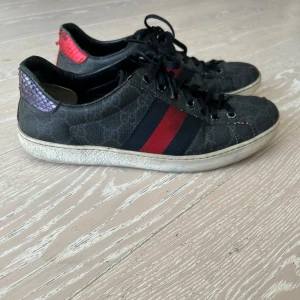 Gucci ace - Svarta Gucci sneakers med GG-monogram, klassiska röd- och blå ränder på sidan och snörning. Baksidan har en röd och en lila metallic detalj i ormskinnsmönster. Snygg och unik design som sticker ut.