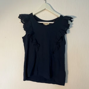 Mörkblå volangtop från Holly & Whyte - Supersöt mörkblå topp från Holly & Whyte i storlek XS med broderade volangdetaljer på axlarna. Ärmlös modell. Ny med prislapp 