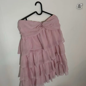 Rosa volangklänning axelbandslös - Supersöt axelbandslös kortklänning i ljusrosa med flera lager volanger och knytdetalj framtill. Klänningen har dragkedja i ryggen och är gjord i ett lätt, luftigt material som ger en romantisk vibe.