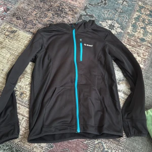 Svart träningsjacka från SOC med huva - Svart träningsjacka från SOC med huva och turkos dragkedja. Jackan har två stora fickor framtill och en liten bröstficka med dragkedja. Tillverkad i mjuk polyester, perfekt för aktiva dagar. Snygg och sportig look med lång ärm och normal passform.