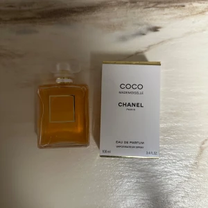Chanel Coco Mademoiselle EdP 100ml - Chanel Coco Mademoiselle Eau de Parfum i en lyxig, rektangulär glasflaska med guldig etikett och vit frostad kork. Parfymen är bärnstensfärgad och kommer i en elegant vit och guldig kartong. Flaskan rymmer 100 ml och utstrålar klassisk och tidlös stil.
