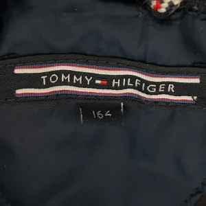 Blå dunjacka från Tommy Hilfiger - Snygg blå dunjacka från Tommy Hilfiger med huva och quiltad design. Jackan är perfekt för kalla dagar och har en klassisk pufferstil som håller dig varm. Passar dig som gillar stilrena och sportiga plagg med en modern vibe.