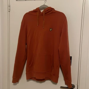 Orange hoodie från Lyle & Scott - Snygg orange hoodie från Lyle & Scott med klassisk gul logga på bröstet. Tröjan har huva med snörning och en stor magficka. Perfekt för dig som gillar en avslappnad och stilren look. Materialet känns mjukt och skönt mot huden.