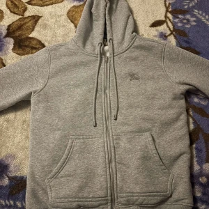 Grå hoodie från Burberry med dragkedja - Snygg grå hoodie från Burberry med klassiskt rutigt foder i huvan och insidan. Tröjan har dragkedja, snörning i huvan och två stora fickor framtill. Tillverkad i mjuk bomullsmix, perfekt för chill dagar. Ikonisk Burberry-logga broderad på bröstet.