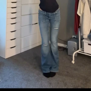 Blå bootcut jeans med slitningar - Säljer ett par snygga blå bootcut jeans med slitningar på knäna och lätt tvättad look. Jeansen har låg midja och klassisk femficksdesign. Perfekta för dig som gillar en avslappnad och trendig stil.