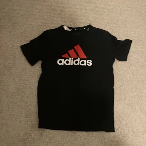 Svart Adidas t-shirt med röd logga - Svart t-shirt från Adidas med stor röd och vit logga på bröstet och en mindre röd logga på ryggen. Klassisk rund hals och korta ärmar. Perfekt för dig som gillar sportig stil och vill ha en enkel men snygg tee med tydlig Adidas-vibe.