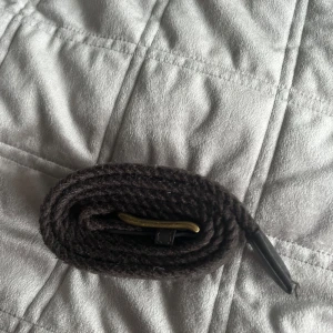 Mörkbrunt stickat bälte med mässingsspänne - Snyggt mörkbrunt bälte i stickat material med klassiskt mässingsfärgat spänne och detaljer i svart skinn. Perfekt accessoar för att lyfta din outfit med en cool och tidlös känsla.