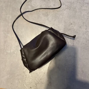 Svart axelväska i skinn - En stilren svart axelväska i mjukt skinn med minimalistisk design. Väskan har en smal axelrem och stängs med en enkel dragsko. Perfekt för dig som gillar en clean look och vill ha med det viktigaste på stan.