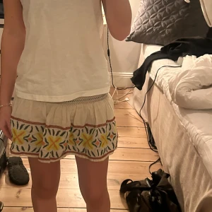 Beige kortkjol med broderi - Superfin beige kortkjol med broderade blommor i gult, orange och grönt. Kjolen har volangkant och bred resår i midjan med dekorativa sömmar. Perfekt för sommarens alla äventyr och ger en bohemisk vibe.