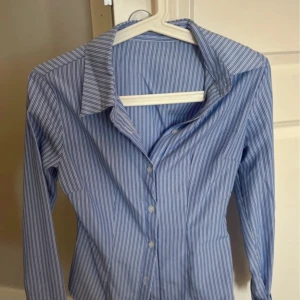 Figursydd randig skjorta  - Snygg blå och vit randig skjorta som är för liten. Skjortan har figurnära passform och knäppning framtill. Vet inte vart den är ifrån. Xxs-xs