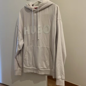 Vit oversized hoodie från HUGO - Säljer en vit hoodie från HUGO med stor logga framtill. Hoodien har huva med dragsko, magficka och en oversized passform. Perfekt för dig som gillar en clean och stilren look. Materialet känns mjukt och skönt mot huden. Äkta!