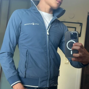 Blå vindjacka från Sail Racing  - •Färg: Marinblå •Storlek: XS och han på bilden är ca 178cm •Modell: Lättviktsjacka med dragkedja framtill •Material: Vind- och vattenavvisande (perfekt för vår, sommar och höst) •Detaljer: Två sidfickor med dragkedja, logga på bröstet, resår vid ärmslut och nederkant med vit rand och innerficka  •Skick: Mycket fint, sparsamt använd  En stilren och funktionell vind jacka från Sail Racing 🧥  Kolla gärna in profilen, det finns flertalet märkeskläder och skor. Mått : Bredd = 44cm, Längd = 59cm