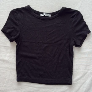 Svart croppad topp från Zara - En svart, enkel och croppad topp från Zara med rund halsringning och korta ärmar. Perfekt att matcha med höga jeans eller kjol för en clean look. Tillverkad i mjuk bomull som känns skön mot huden.