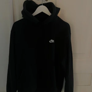 Svart hoodie från Nike med huva - Svart hoodie från Nike med klassisk vit logga på bröstet. Tröjan har huva och långa ärmar, perfekt för en avslappnad stil. Mjuk och bekväm, passar till många tillfällen.