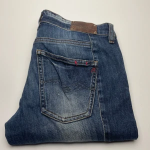 Blå jeans Replay Radula W27 L30 - Snygga blå jeans från Replay, modell Radula. Klassisk femficksdesign med slitningar och kontrastsömmar. Jeansen har röda detaljer vid fickorna och är tillverkade i bomull med en skön jeanskänsla. Perfekta för en avslappnad och cool stil.