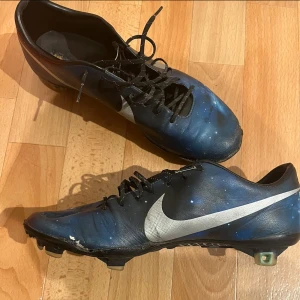 Blåa Nike fotbollsskor med silverlogga - Säljer ett par blåa Nike fotbollsskor med silverfärgad swoosh på sidan. Skorna har snörning och platt sula med dobbar för bra grepp på planen. Materialet är syntet och designen har en cool galax-liknande effekt.