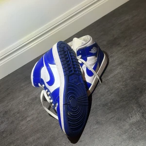 Nike Air Jordan 1 blå och vita sneakers - Säljer ett par Nike Air Jordan 1 sneakers i blått och vitt skinn. Klassisk high-top modell med vit bas, blå detaljer och vit sula. Ikoniska Jordan Wings-loggan på sidan och perforerad tå för extra stil. Perfekta för dig som vill sticka ut med en tidlös streetwear vibe. Utan box