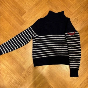 Stickad polotröja från Zadig & Voltaire - Mörkblå och vit randig polotröja från Zadig & Voltaire med broderad röd logga på ena ärmen. Tröjan är ribbstickad, har lång ärm och hög polokrage. Perfekt för dig som gillar en cool och klassisk vibe.