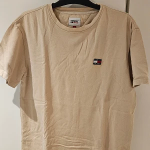 Beige t-shirt från Tommy Jeans - Snygg beige t-shirt från Tommy Jeans med liten broderad logga på bröstet. Klassisk rund hals och korta ärmar. Tillverkad i 100% bomull för en skön och avslappnad känsla. Perfekt till jeans eller shorts för en clean look.