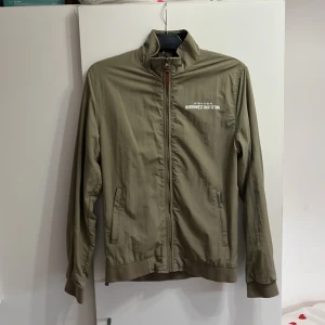 Olivgrön vindjacka från Smog - Säljer en olivgrön vindjacka från Smog med dragkedja och ståkrage. Jackan har två fickor framtill och tryckt text på bröstet. Insidan har ett snyggt bladmönster i brunt och mörkblått. Perfekt för dig som vill ha en stilren och lätt jacka.