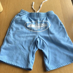 Blå shorts från corteiz - Snygga ljusblå shorts från Eytys med vitt tryck av ett skepp på framsidan. Modellen har elastisk midja med vit snörning, sidofickor och en bakficka med lock. Mjukt material i bomull.