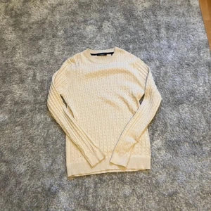 Beige stickad tröja från Jack & Jones - Säljer en stilren beige stickad tröja från Jack & Jones. Tröjan har ett klassiskt flätmönster och långa ärmar, perfekt för kyligare dagar. Den är mjuk och bekväm, idealisk för både vardag och avslappnade tillfällen.