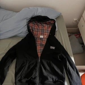 Svart Burberry hoodiejacka med rutig insida - Svart hoodiejacka från Burberry med dragkedja och klassiskt rutigt foder i beige, rött och svart. Jackan har huva med snörning, två fickor fram och Burberry London England-logga på bröstet. Perfekt för dig som vill ha en stilren streetlook med lyxig touch.