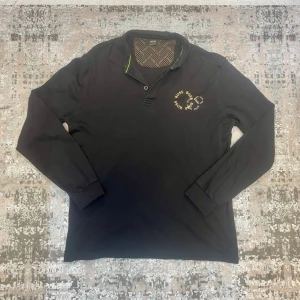 Svart långärmad pikétröja från BOSS - Svart långärmad pikétröja från BOSS med tryckt logga i gult och vitt på bröstet och en snygg detalj på ärmen. Kragen har två knappar och insidan har ett mönstrat tyg. Perfekt för dig som gillar stilrena och klassiska plagg.