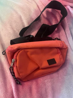 Rosa axelväska från 157 - Säljer en snygg rosa axelväska från 157 med svart justerbar rem och praktiska dragkedjor. Väskan har en stilren design och är perfekt för att bära dina viktigaste grejer. Tillverkad i slitstark polyester och har en liten logga framtill.