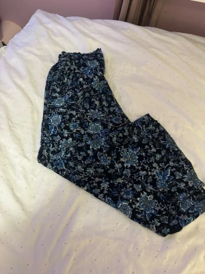Blommiga byxor från H&M, strl 38 - Säljer ett par flowiga byxor från H&M i storlek 38. Byxorna är mörkblå med ett snyggt blommigt mönster i blå och vita toner. De har resår i midjan och är gjorda i ett mjukt, tunt material som känns skönt mot huden.