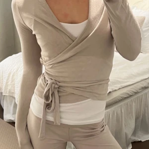 Beige omlottopp med knyt - Snygg beige omlottopp med långa ärmar och knyt i midjan. Toppen har en v-ringad design och är gjord i mjukt material som sitter skönt mot kroppen. Perfekt för dig som gillar stilrena och enkla plagg med en twist. Storlek XS-S