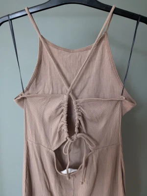 Beige ribbad kort klänning med snörning - Säljer en beige ribbad kortklänning med tunna axelband och snygg snörningsdetalj framtill. Klänningen har en cut-out under bysten och är tillverkad i mjukt, stretchigt material. Perfekt för varma dagar och enkel att styla med sneakers eller sandaler.