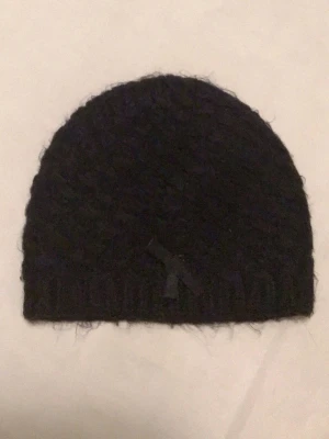 Svart stickad mössa med rosett - Mysig svart stickad mössa med liten rosett framtill. Mössan har en enkel, klassisk form och är tillverkad i ett fluffigt material som ger en soft känsla. Perfekt för kalla dagar när du vill hålla värmen och ändå se stilren ut.