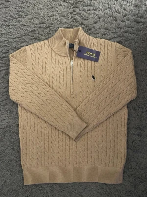 Beige kabelstickad tröja Polo Ralph Lauren - Säljer en beige kabelstickad tröja från Polo Ralph Lauren med half zip och hög krage. Tröjan har lång ärm, ribbade muddar och den klassiska broderade loggan på bröstet. Perfekt för dig som gillar stilrena och tidlösa plagg.