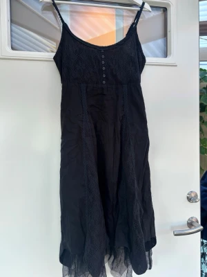 Svart spetsklänning från Cream - Supersnygg svart långklänning från Cream med tunna axelband, spetsdetaljer och knappar framtill. Klänningen har ett lager av transparent tyg längst ner och vackra spetsinslag längs hela plagget. Perfekt för dig som gillar en bohemisk och romantisk stil.