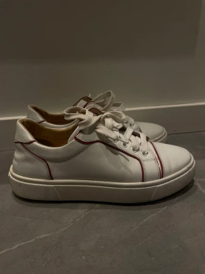 Vita sneakers från Louboutin - Säljer ett par vita sneakers från Louboutin med röda detaljer och klassisk röd sula. Skorna har rund tå, snörning och är tillverkad i skinn. Köpta på vestiare collective och finns äkthetsbevis ifrån dem (se bild 7). Storlek 38,5.