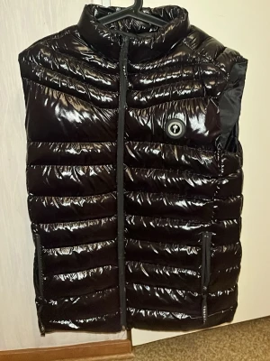 Svart dunväst från Trapstar - Säljer en svart, glansig dunväst från Trapstar med broderad logga på bröstet och stor Trapstar-text på ryggen. Västen har dragkedja framtill och två fickor med dragkedja. Perfekt för lager-på-lager och streetwear-vibe.