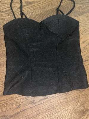 Glittrig svart korsettopp - Snygg svart korsettopp med tunna axelband och glittrigt tyg. Toppen har markerade kupor och en croppad passform som ger en trendig siluett. Stoppen har justerbara band! Använd 1 gång 