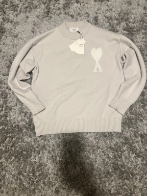 Ljusgrå kashmirtröja från Ami - Supersnygg ljusgrå tröja från Ami med ikoniskt hjärta och A på bröstet. Tillverkad i mjuk kashmir och ullblandning, har ribbade muddar och rund hals. Perfekt för dig som gillar stilrena och exklusiva plagg.