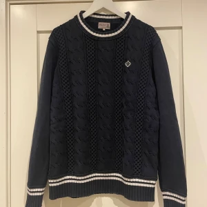 Morris svart stickad tröja - Säljer en svart stickad tröja från Morris med kabelstickat mönster framtill och vita ränder på krage, ärmslut och nederkant. Tröjan har rund hals och en broderad logga på bröstet. Perfekt för dig som gillar klassisk stil.