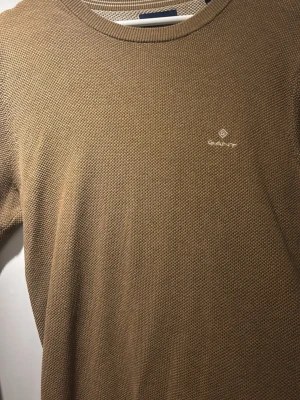 Beige stickad tröja från GANT - Säljer en beige stickad tröja från GANT i storlek M. Tröjan har rund halsringning, diskret GANT-logga broderad på bröstet och ribbade muddar vid ärmslut och hals. Perfekt för lager på lager under hela året. 